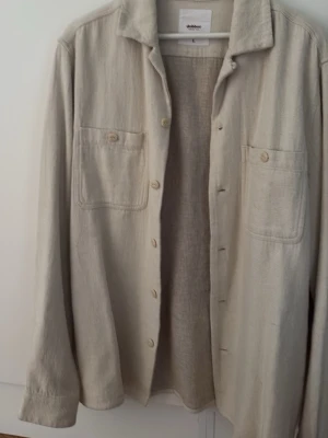 Beige overshirt från Dobber - Stilren beige overshirt från Dobber i storlek L. Skjortan har två bröstfickor med knapp, klassisk krage och knäppning framtill. Tillverkad i ett mjukt, vävt material som ger en avslappnad look. Perfekt att slänga över en t-shirt för en cool lager-på-lager-stil.