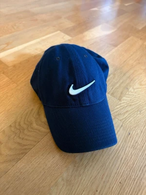 Mörkblå Nike keps med broderad logo - Snygg mörkblå keps från Nike med den klassiska vita swoosh-loggan broderad framtill. Justerbar rem baktill för perfekt passform. Tillverkad i bomullstyg som andas och känns bekvämt på huvudet. Perfekt accessoar för en sportig och avslappnad stil.
