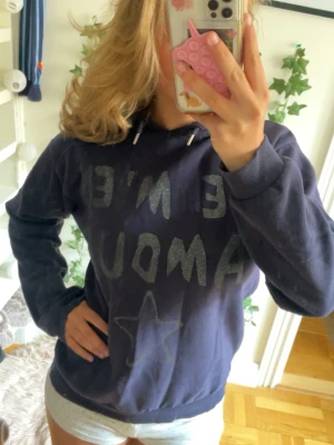 Mörkblå hoodie med text och stjärna - Säljer en mörkblå hoodie med huva och snören. På framsidan står det 'JE M'EN AMOUR' i glittrig text och en stjärna under. Hoodien har ribbade muddar vid ärmar och nederkant. Perfekt för en chill och avslappnad stil.