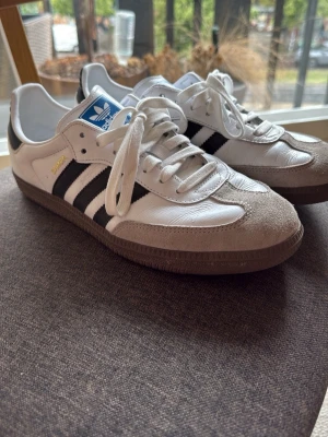 Adidas Samba OG vita sneakers - Klassiska Adidas Samba OG sneakers i vitt läder med svarta ränder och grå mockadetaljer på tån. Skorna har vit sula, blå Adidas-logga på plösen och gul Samba-text på sidan. Perfekta för en sportig och retro look.