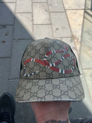 Gucci keps med orm och GG-mönster - Säljer en snygg Gucci keps med klassiskt GG-mönster i beige och grått. Framtill finns en röd och vit orm tryckt över hela kepsen. Baksidan är i svart mesh med justerbart band i grönt och rött. Perfekt för dig som gillar streetwear och exklusiva accessoarer.