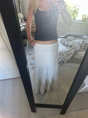 Vit långkjol med smock i midjan - Säljer en vit långkjol med smockdetalj i midjan och lätt volang nedtill. Kjolen har en luftig och flowy passform, perfekt för varma dagar. Materialet känns mjukt och svalt, troligen bomull eller linne.