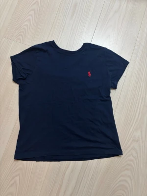 Marinblå t-shirt från Ralph Lauren - Klassisk marinblå t-shirt från Ralph Lauren med röd broderad logga på bröstet. T-shirten har rund halsringning och korta ärmar. Tillverkad i mjuk bomull för en skön känsla och enkel stil.