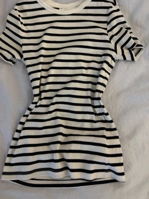 Randig ribbad t-shirt från H&M - Säljer en vit och svart ribbad t-shirt från H&M med klassiskt randigt mönster. T-shirten har korta ärmar och rund halsringning. Perfekt för en enkel och stilren look.