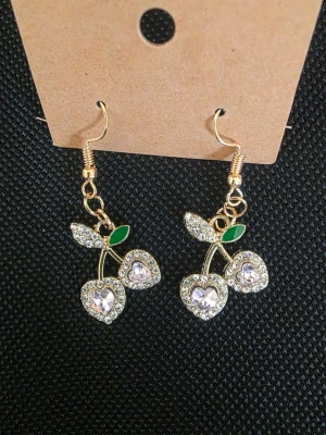 Handgjorda Guldörhängen med körsbär och strass - Handgjorda Snygga hängande örhängen i guldfärg med körsbärsformade hängen. Körsbären är dekorerade med glittrande strass och hjärtformade stenar, samt små gröna blad. Perfekta för dig som gillar statement-smycken med lekfull design.