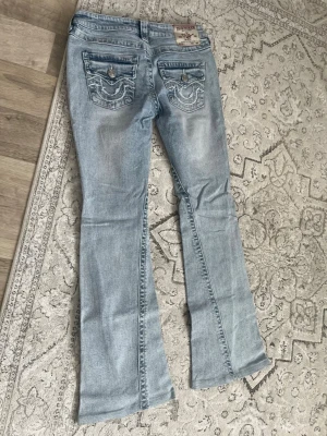 Ljusblå bootcut jeans True Religion - Säljer ett par ljusblå bootcut jeans från True Religion med snygga detaljerade bakfickor och kontrastsömmar. Jeansen har låg midja och klassisk femficksdesign. Har sytt om längden hos skräddare men kändes lite löst i midjan om man är jätte smal som mig, det är därför jag säljer den.  Det är nytt utan prislapp har aldrig använt den 