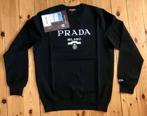 Svart sweatshirt från Prada - Stilren svart sweatshirt från Prada med vit logotyp och texten 'PRADA MILANO' på bröstet. Tröjan har rund halsringning, långa ärmar och ribbade muddar vid ärmslut och nederkant. Perfekt för en trendig och avslappnad look.