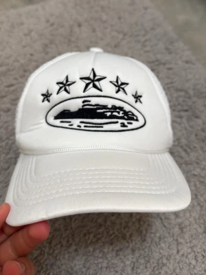 Vit meshkeps med broderade stjärnor - Snygg vit keps med svart broderat motiv framtill, fem stjärnor och ett ovalt märke. Baksidan är i mesh för extra ventilation och har justerbar snapback. Perfekt för en streetwear-look.