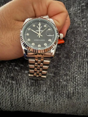 Silverfärgad klocka från DEEFRUN - Stilren klocka från DEEFRUN med silverfärgat metallarmband och svart urtavla. Urtavlan har markerade index med stenar och datumvisning. Klassisk rund form och räfflad kant runt glaset ger en exklusiv känsla.