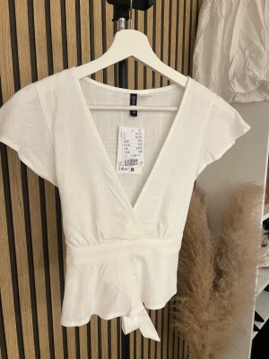 Vit omlottopp från H&M - Snygg vit omlottopp från Divided H&M med korta ärmar och knyt i midjan. Toppen har en djup v-ringning och är tillverkad i ett lätt bomullsliknande material. Perfekt för en fräsch och enkel look.