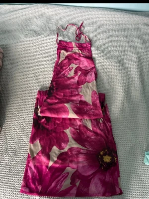 Vintage Blommig långklänning - Säljer en långklänning med stora rosa blommor på beige botten. Klänningen har tunna axelband som knyts i nacken och en figurnära passform. Perfekt för sommarens alla tillfällen!
