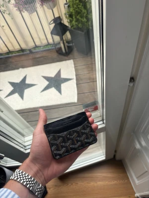 Svart plånka från GO…. - Snyggt svart kortfodral med det klassiska mönstret i vitt, brunt och grått. Fodralet har flera fack för kort och är gjort i en kombination av canvas och läder. Perfekt för dig som vill ha något stilrent och exklusivt i fickan. Obs, den är använd.