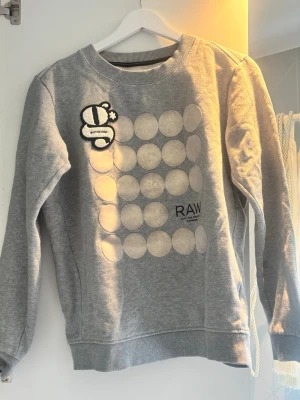 Grå sweatshirt från G-Star RAW - Säljer en grå sweatshirt från G-Star RAW med rund hals och ribbade muddar. Framsidan har ett coolt cirkelmönster i vitt och en broderad patch med G-logga på bröstet. Perfekt för en avslappnad streetwear-look.