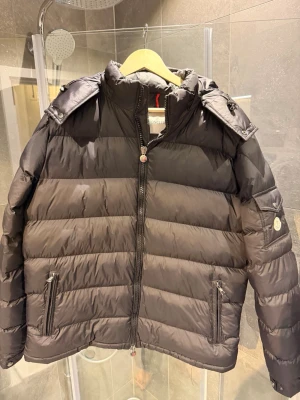 Svart dunjacka från Moncler - Snygg svart dunjacka från Moncler med avtagbar huva, två dragkedjefickor fram och en ficka med logotyp på ärmen. Jackan är i bra skick, och det finns inga defeker.  