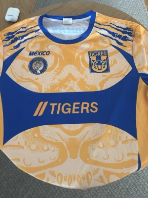 Tigres UANL matchtröja gul/blå - Snygg gul och blå matchtröja från Tigres UANL med tryckta loggor, tiger-motiv och texten 'TIGERS' på bröstet. På ryggen står det 'TIGERS LET'S GO'. Perfekt för dig som älskar mexikansk fotboll och vill sticka ut på läktaren eller träningen.