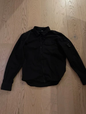 Svart overshirt från C.P. Company - Säljer en svart overshirt från C.P. Company med klassisk krage, bröstficka och dragkedja framtill. Ärmen har den ikoniska linsen på fickan, vilket ger en unik touch. Overshirten är tillverkad i ett lätt material och har en clean, modern look.
