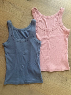 Ribbad blå och rosa linne Xs - Två ribbade linnen i blått och ljusrosa. Båda har rundad halsringning och är ärmlösa, perfekta för sommaren. Snygga att matcha med jeans eller kjol för en enkel och trendig look.