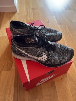 Nike Flyknit Racer svart/vit - Nike Flyknit Racer sneakers i svart och vitt med stickad ovandel och klassisk vit swoosh på sidan. Lätta och ventilerande med svart snörning och låg profil. Sulan är vit med svarta detaljer. Perfekta för dig som gillar stilrena och sportiga sneakers.