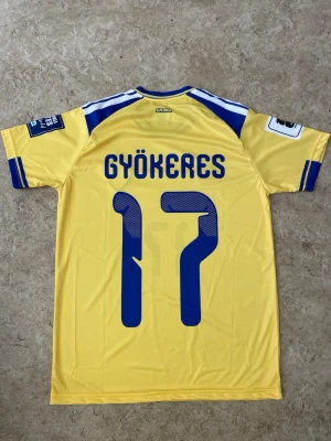 Svensk landslagströja Gyökeres 17 - Gul och blå svensk landslagströja från Adidas med Gyökeres och nummer 17 på ryggen. Tröjan har korta ärmar, blå detaljer på axlar och ärmslut samt Sveriges landslagsemblem på bröstet. Tillverkad i lätt och ventilerande material, perfekt för fotboll.