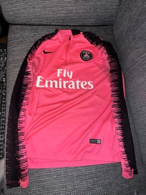 Rosa PSG träningströja från Nike - Säljer en snygg rosa långärmad träningströja från PSG med svart mönster längs ärmarna och vit Fly Emirates-logga på bröstet. Tröjan har halv dragkedja och klubbmärke på vänster sida. Perfekt för fotbollsträning eller gymmet.