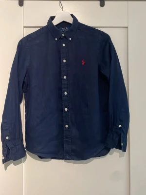 Marinblå skjorta från Polo Ralph Lauren M10-12 - Klassisk marinblå skjorta från Polo Ralph Lauren med knappkrage och vita knappar. Skjortan har lång ärm och den ikoniska röda logotypen broderad på bröstet. Tillverkad i mjuk bomull för en bekväm känsla.