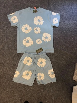Denim Tears blå set - Säljer ett ljusblått set från Denim Tears med matchande t-shirt och shorts. Båda plaggen har vita blommönster tryckta över hela ytan. Setet är gjort i mjukt bomullsmaterial och har en avslappnad passform. Perfekt för dig som gillar streetwear och unika prints.Finns andra färger. Finns storlek S och M
