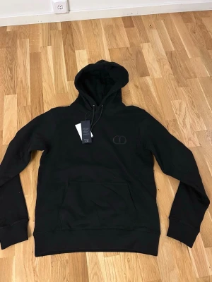 Svart hoodie från dior  - Säljer en svart hoodie från dior med klassisk huva och dragsnören. Tröjan har en stor magficka och diskret logga på bröstet. Materialet är mjukt och bekvämt, perfekt för en avslappnad stil.