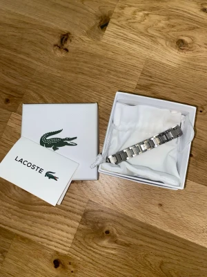 Silverfärgat armband från Lacoste - Snyggt silverfärgat armband från Lacoste i metall med stilrena länkar och diskret Lacoste-logga. Kommer i original ask och tygpåse med krokodiltryck. Perfekt accessoar för dig som gillar klassisk och sportig stil.