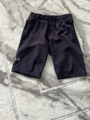 Svarta träningsshorts från Under Armour - Snygga svarta träningsshorts från Under Armour med elastisk midja och diskret logga på benet. Shortsen har en praktisk ficka med dragkedja bak och är gjorda i ett lätt syntetmaterial som andas. Perfekta för dig som gillar att röra på dig.
