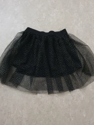 Tyllkjol - Snygg svart kortkjol i tyll med små glittriga guldprickar. Kjolen har ett elastiskt midjeband och flera lager tyll som ger extra volym. Perfekt för dig som vill sticka ut lite extra med en lekfull och trendig look.