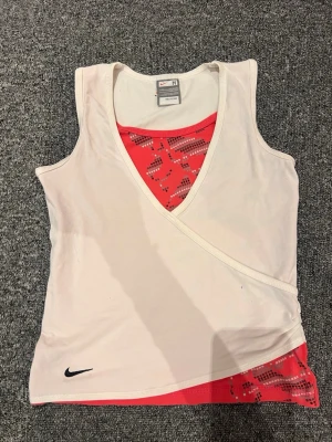 Nike sportlinne med omlott och mönster - Snyggt sportlinne från Nike i vitt med omlott-design och ett rött innerlager med grafiskt mönster. Linnet har djup V-ringning och ärmlös passform, perfekt för träning. Tillverkat i ett mjukt och stretchigt material för maximal komfort.