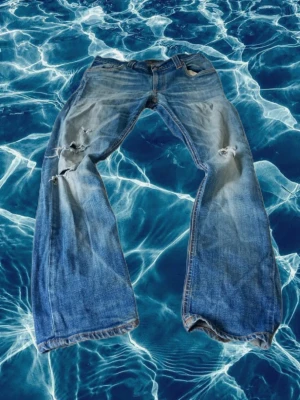 Blå slitna jeans från nudie - Ett par blå jeans med slitningar och hål på båda benen. Jeansen har klassisk femficksmodell och raka ben. Materialet är denim i bomull och de har en avslappnad, vintage look. De är Nudie jeans. 29/32