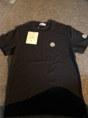 Svart Moncler t-shirt med logga - Snygg svart t-shirt från Moncler med klassisk logga på bröstet och ärmen. T-shirten har rund hals och korta ärmar, tillverkad i mjuk bomull för skön passform. Perfekt till jeans eller shorts för en clean och stilren look.