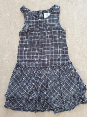 Klänning - Säljer en grå ärmlös klänning från H&M i rutigt mönster. Klänningen har en avslappnad passform med volangdetaljer nedtill och är tillverkad i mjuk bomull. Perfekt för dig som gillar en enkel men snygg stil.