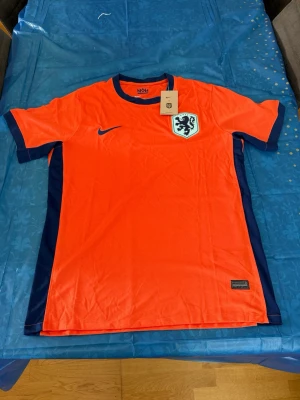 Holland Nike fotbollströja orange - Snygg orange fotbollströja från Nike med Nederländernas landslagsemblem på bröstet och mörkblå detaljer vid ärmar och sidor. Tillverkad i lätt och ventilerande material, perfekt för träning eller match. Klassisk passform och rund hals.