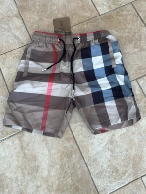 Burberry shorts/badbyxor - Påse ingår. Säljer ett par klassiska Burberry shorts med rutigt mönster i beige, brun, blå, vit och röd. Shortsen har elastisk midja med svart snörning och är perfekta för sommaren. Ikonisk design som sticker ut och passar till många olika stilar. 