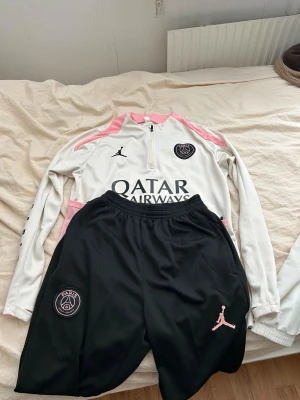 PSG träningsset vit/rosa/svart - Säljer ett PSG träningsset med långärmad vit tröja med rosa detaljer och svart logga, samt svarta byxor med rosa Jordan-logga och PSG-märke. Tröjan har halv dragkedja och tryck från Qatar Airways. Perfekt för fotbollsträning.