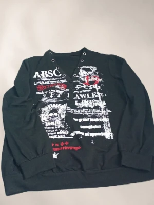 Svart hoodie med tryck och nitar - Cool svart hoodie med tryck i vitt och rött framtill, detaljer som dödskalle och text. Unik design med nitar och knappdetaljer vid halsen. Mjuk bomullskänsla och långa ärmar. Perfekt för dig som gillar alternativ stil och streetwear.
