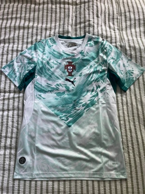 Portugal fotbollströja Puma grön/vit - Snygg Portugal fotbollströja från Puma i vitt och grönt med vågigt mönster. Tröjan har korta ärmar, rund hals och Portugal-emblemet på bröstet. Tillverkad i lätt funktionsmaterial med DryCell-teknologi för bra ventilation.
