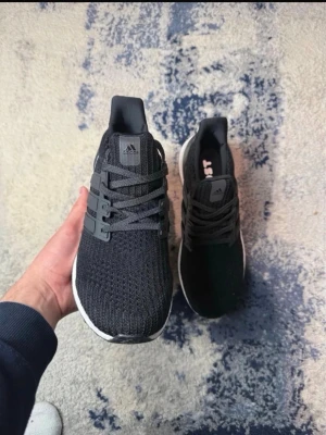 Svarta Adidas Ultraboost sneakers - Säljer ett par svarta Adidas Ultraboost sneakers med stickad ovandel och vita Boost-sulor. Skorna har klassiska tre ränder på sidan, svart hälkappa och grå snörning. Perfekta för dig som vill ha både stil och komfort i vardagen.