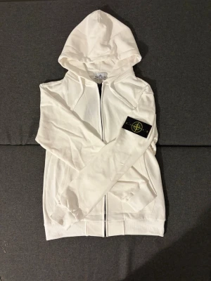 Vit hoodie från Stone Island - Snygg vit hoodie från Stone Island med dragkedja framtill och klassisk logotyp-patch på vänster ärm. Hoodien har huva, ribbade muddar och är tillverkad i mjukt bomullsmaterial. Perfekt för en clean och stilren streetwear-look.