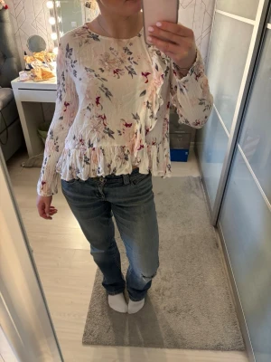 Blommig volangblus i ljusa toner - Söt blus med volangkant och blommigt mönster i rosa, rött och blått på ljus botten. Långärmad modell med rund halsringning och lätt rynkade detaljer. Perfekt för dig som gillar romantisk och feminin stil.