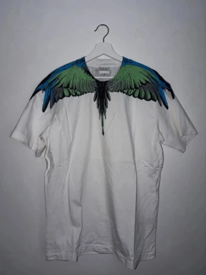Marcelo Burlon  - Säljer en vit t-shirt från Marcelo Burlon med ikoniskt vingtryck i grönt, blått och svart över axlar och bröst. T-shirten har normal passform, korta ärmar och är tillverkad i mjuk bomull. Perfekt för dig som vill sticka ut med en unik design.