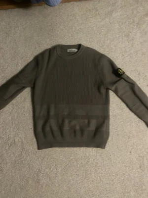 Grå stickad tröja Stone Island - Snygg grå stickad tröja från Stone Island med rund halsringning och ribbade muddar. Tröjan har en ikonisk patch med logga på vänster ärm och är tillverkad i ett tjockare, strukturerat material. Perfekt för dig som gillar streetwear och stilrena plagg.