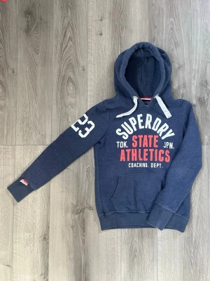 Blå hoodie från Superdry Athletics - Mörkblå hoodie från Superdry med vit och röd text på bröstet och siffran 23 på ärmen🤗 Passar nån med storlek S-Xs🤩 Priset ej hugget i sten därför kan det diskuteras😇