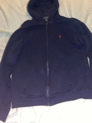 Mörkblå hoodie från Polo Ralph Lauren - Mörkblå hoodie med dragkedja från Polo Ralph Lauren. Klassisk design med huva, kängurufickor och den ikoniska röda logotypen broderad på bröstet. Tillverkad i mjukt bomullsmaterial som är skönt att ha på sig. Perfekt för en avslappnad och stilren look.