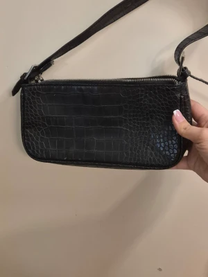 Svart axelväska i krokopräglat skinn - Snygg svart handväska med axelrem och croco-mönstrad yta. Väskan har en rak, avlång form och stängs med dragkedja upptill. Perfekt för dig som gillar trendiga accessoarer med lite edge.