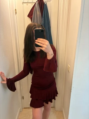 Vinröd volangklänning med långa ärmar - Säljer en vinröd kort klänning med långa trumpetärmar och volangdetaljer vid kjolen. Klänningen har en figurnära passform och är tillverkad i ett mjukt, stretchigt material som känns bekvämt mot huden. Perfekt för dig som vill sticka ut med en elegant look.