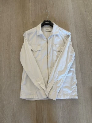 Vit overshirt från These Glory Days - Stilren vit overshirt från These Glory Days med dragkedja framtill och två bröstfickor med lock. Overshirten har klassisk krage och långa ärmar med knapp vid ärmslut. Perfekt att bära som lager över t-shirt. Materialet känns mjukt och lätt.