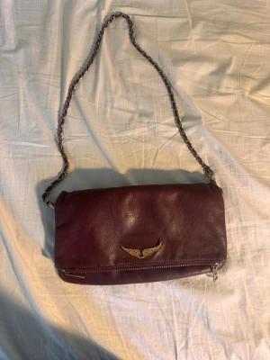 Vinröd Zadig väska med gulddetalj - Snygg vinröd axelväska i mjukt läder med guldfärgad kedjerem och en cool guldfärgad vingdetalj på framsidan. Väskan har dragkedja längst ner och en stilren, enkel design som passar till det mesta. Äkta Zadig köpt på Plick.